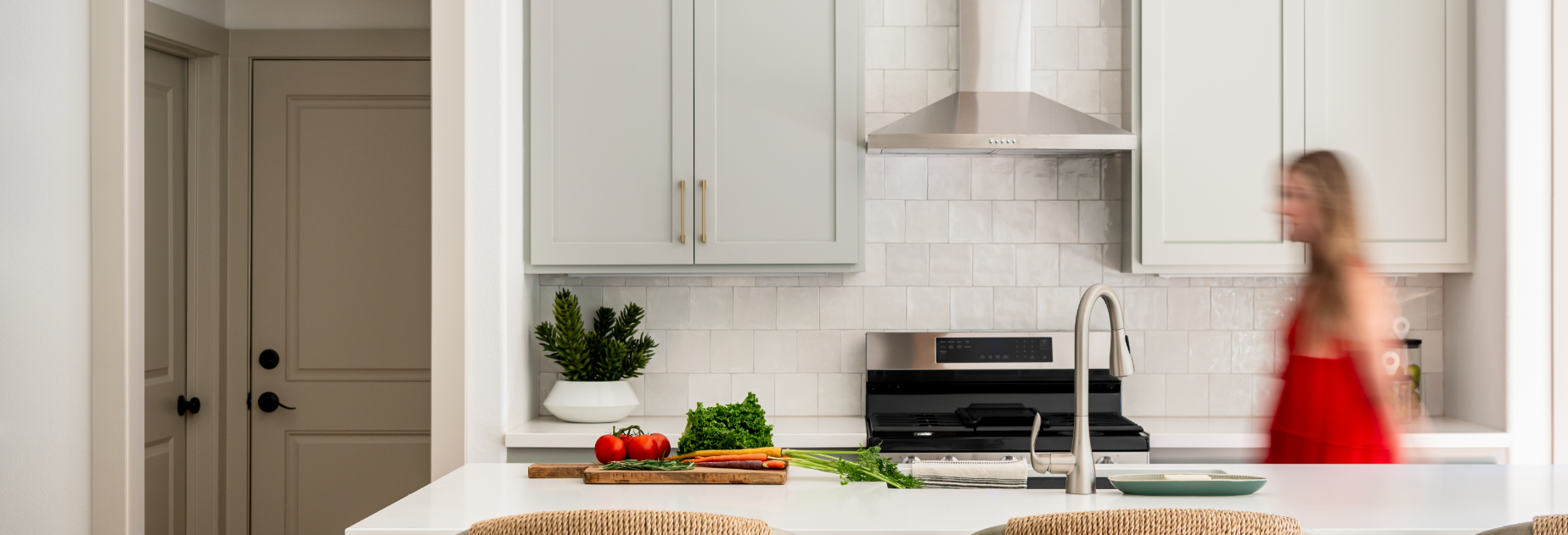 A woman walking in a white kitchen with a sink and a stove.