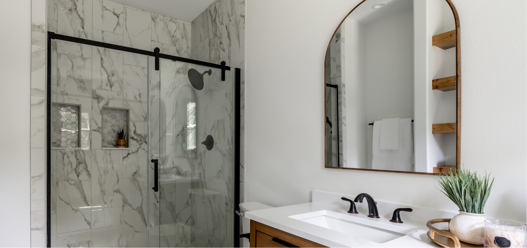 A  bathroom with a glass shower, sink and arched mirror